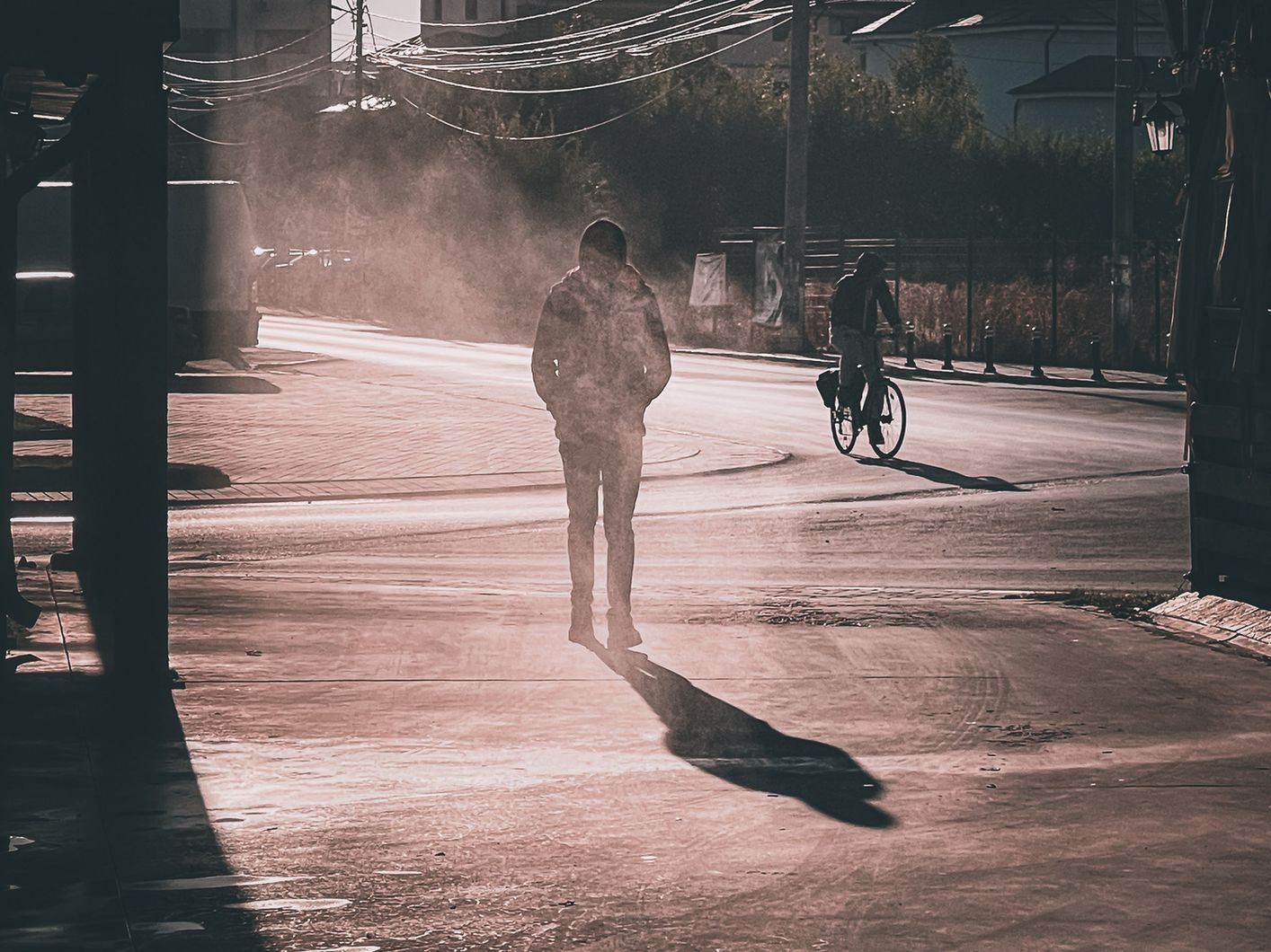 A lone figure standing on a hazy, smoke-filled street at golden hour as a cyclist passes.
